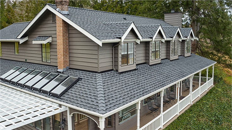 A gray-shingled house with white trim and solar panels on the roof, surrounded by trees.