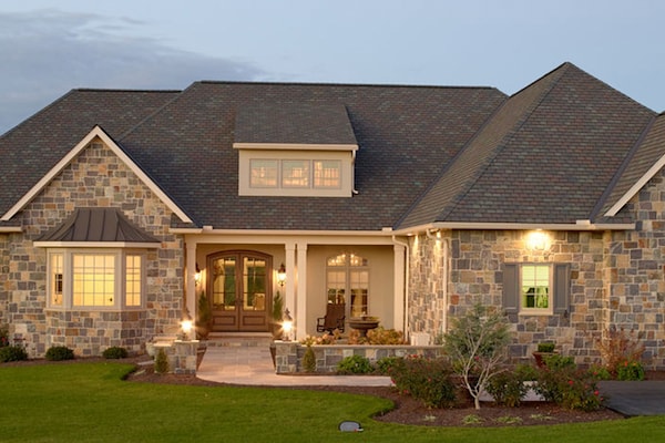 A large stone mansion with warm lighting illuminates its entrance and windows against a twilight sky.