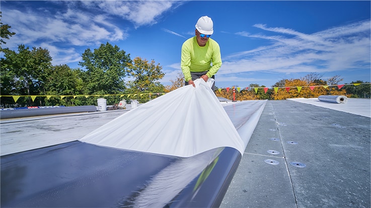 A construction worker in a hard hat and safety vest unrolls a white roofing membrane on a flat roof under a bright blue sky.