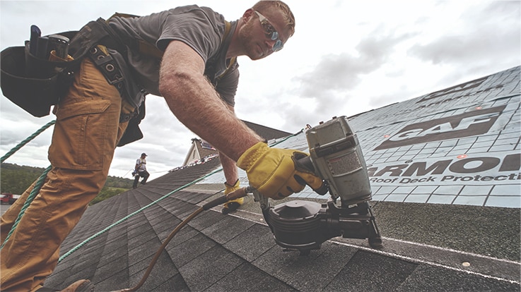 A roofer uses a nail gun to install asphalt shingles on a sloped roof under an overcast sky.