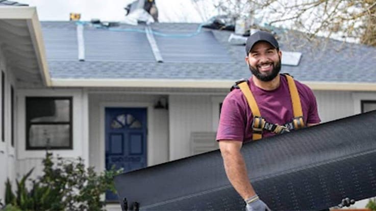 A smiling technician carries a solar panel in front of a house with installed panels on the roof.