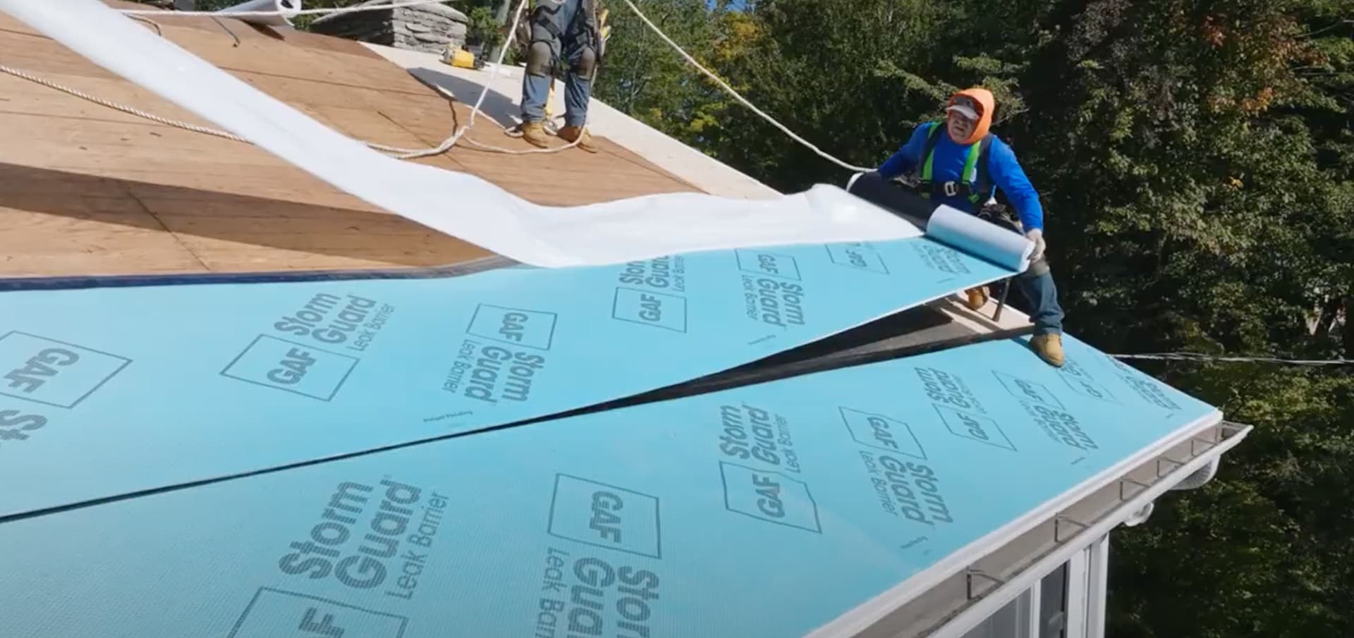 A worker rolls out blue GAF Storm Guard leak barrier on a sloped roof under bright sunlight.